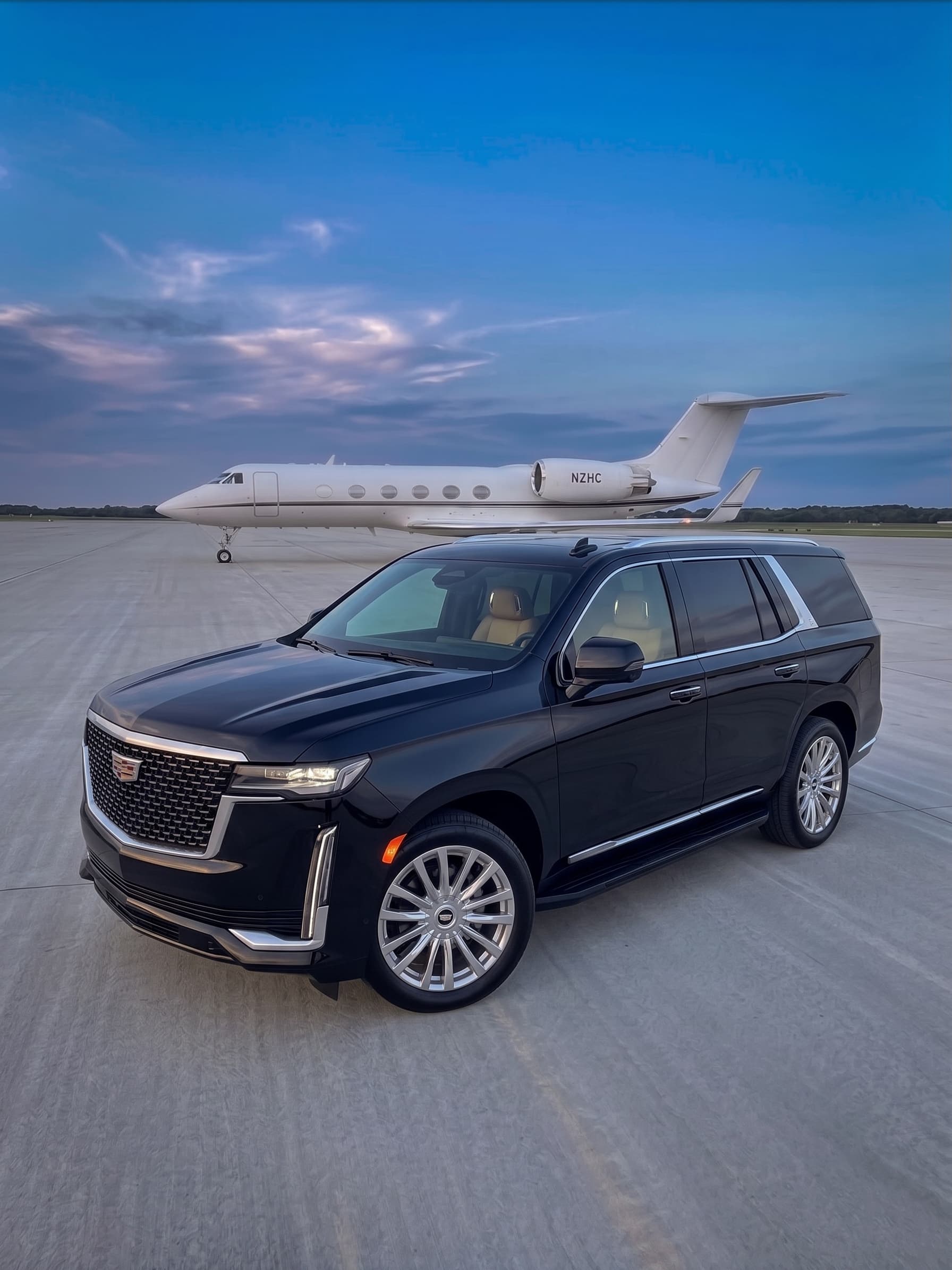 A MUVEH Cadillac Escalade on a private aviation tarmac at dusk, with a jet behind it
