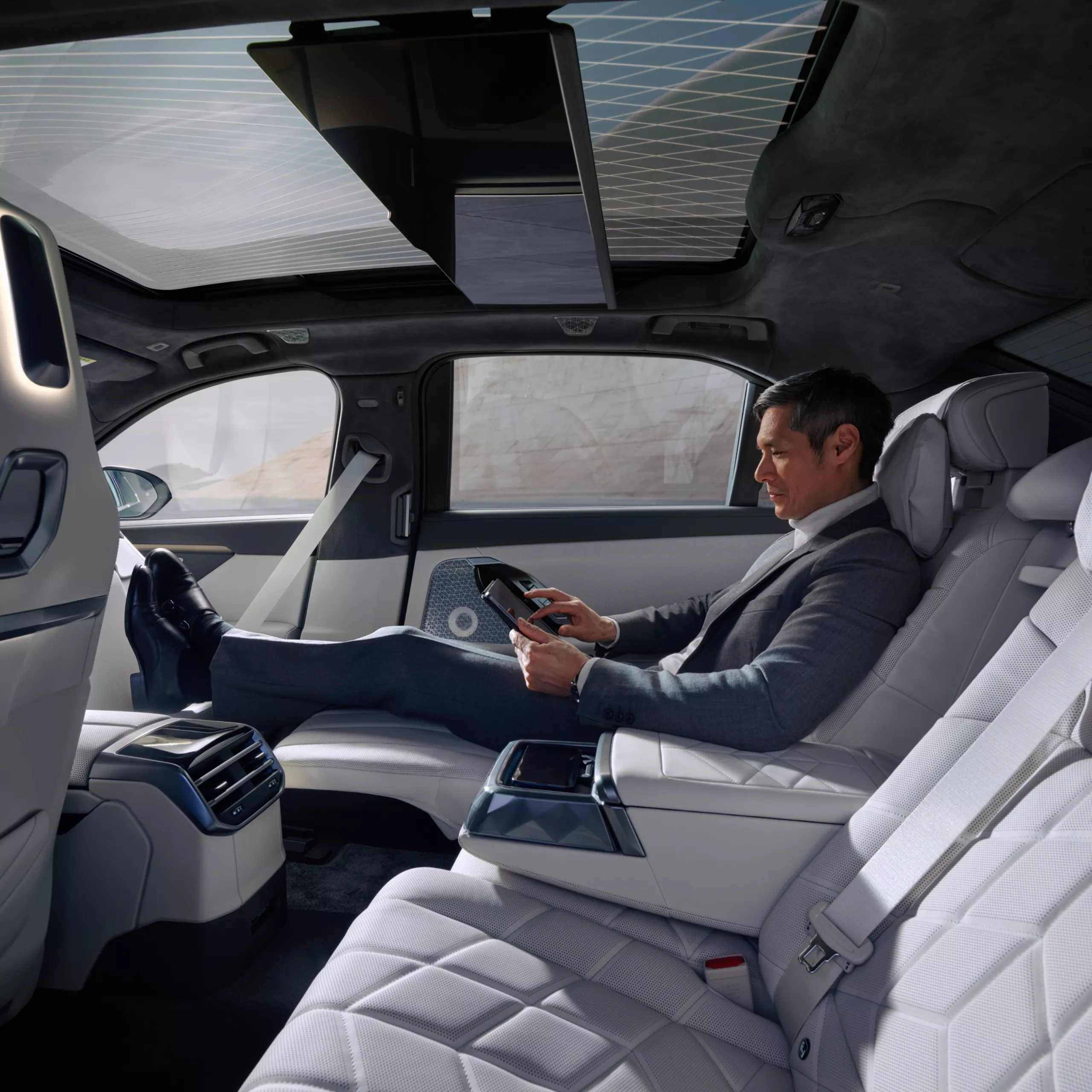A passenger reclined in the rear seat of a BMW 7 Series, tablet in hand beneath a panoramic roof