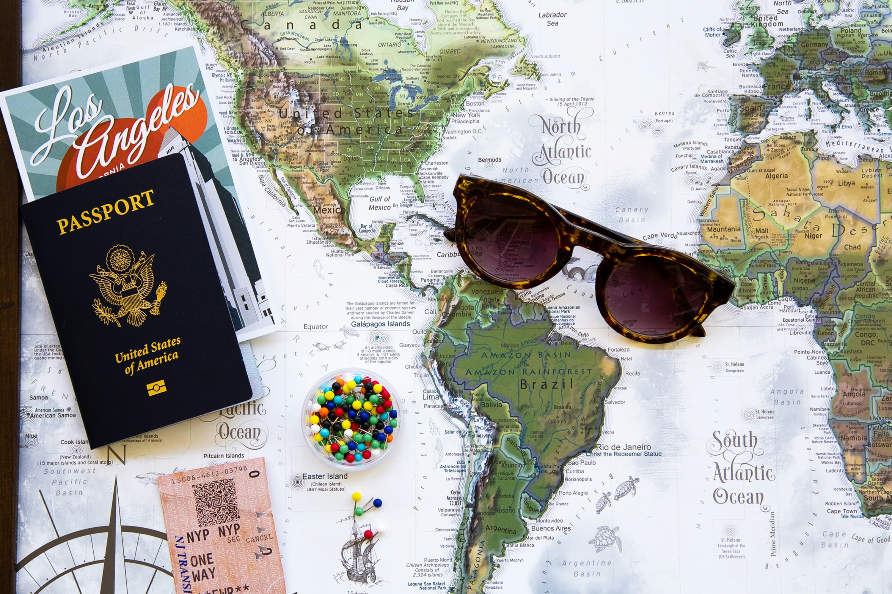 A world map laid out with a US passport, sunglasses, a boarding pass and a handful of coloured pins