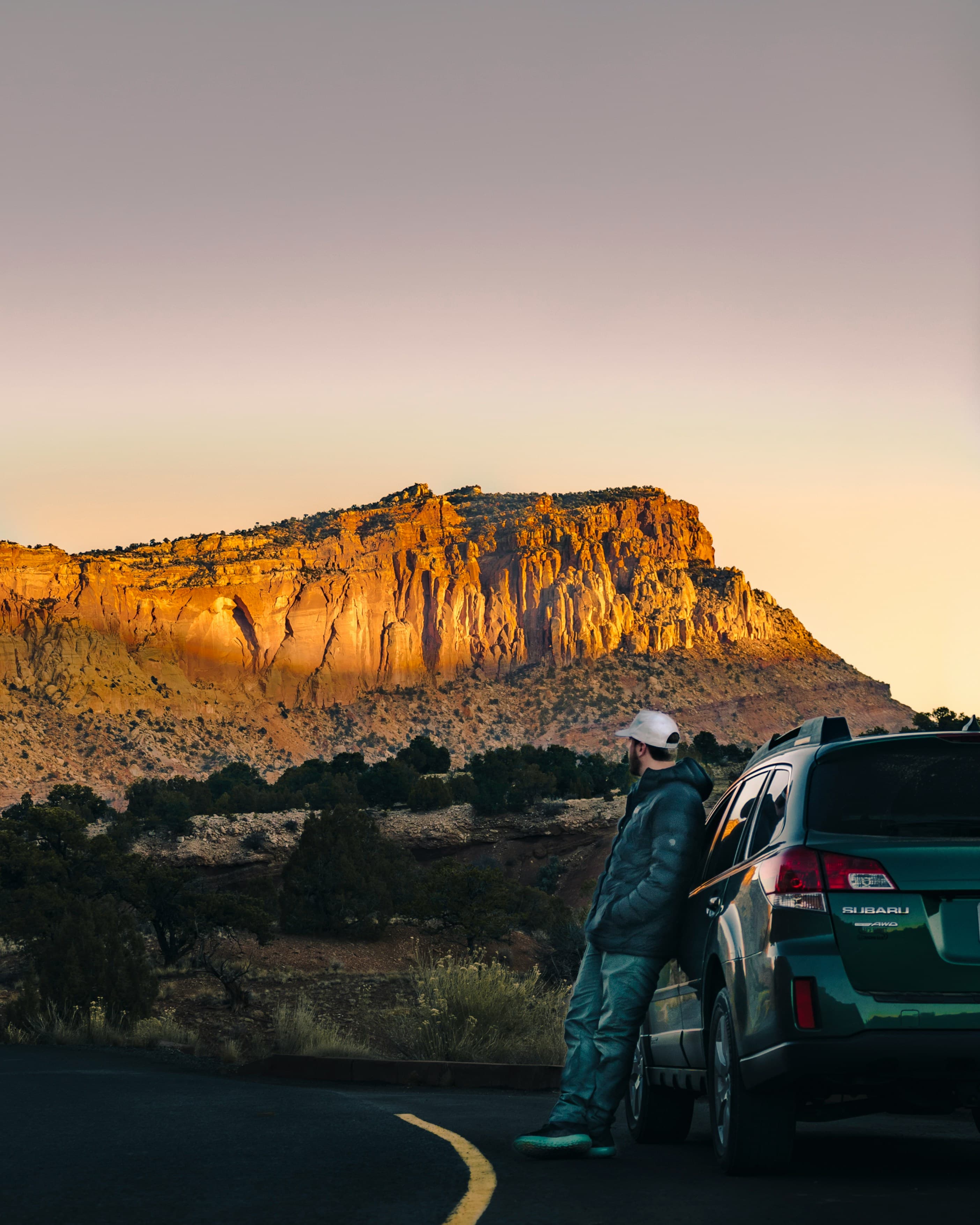 A traveller leaning against a car at dusk, looking out at a glowing red-rock cliff
