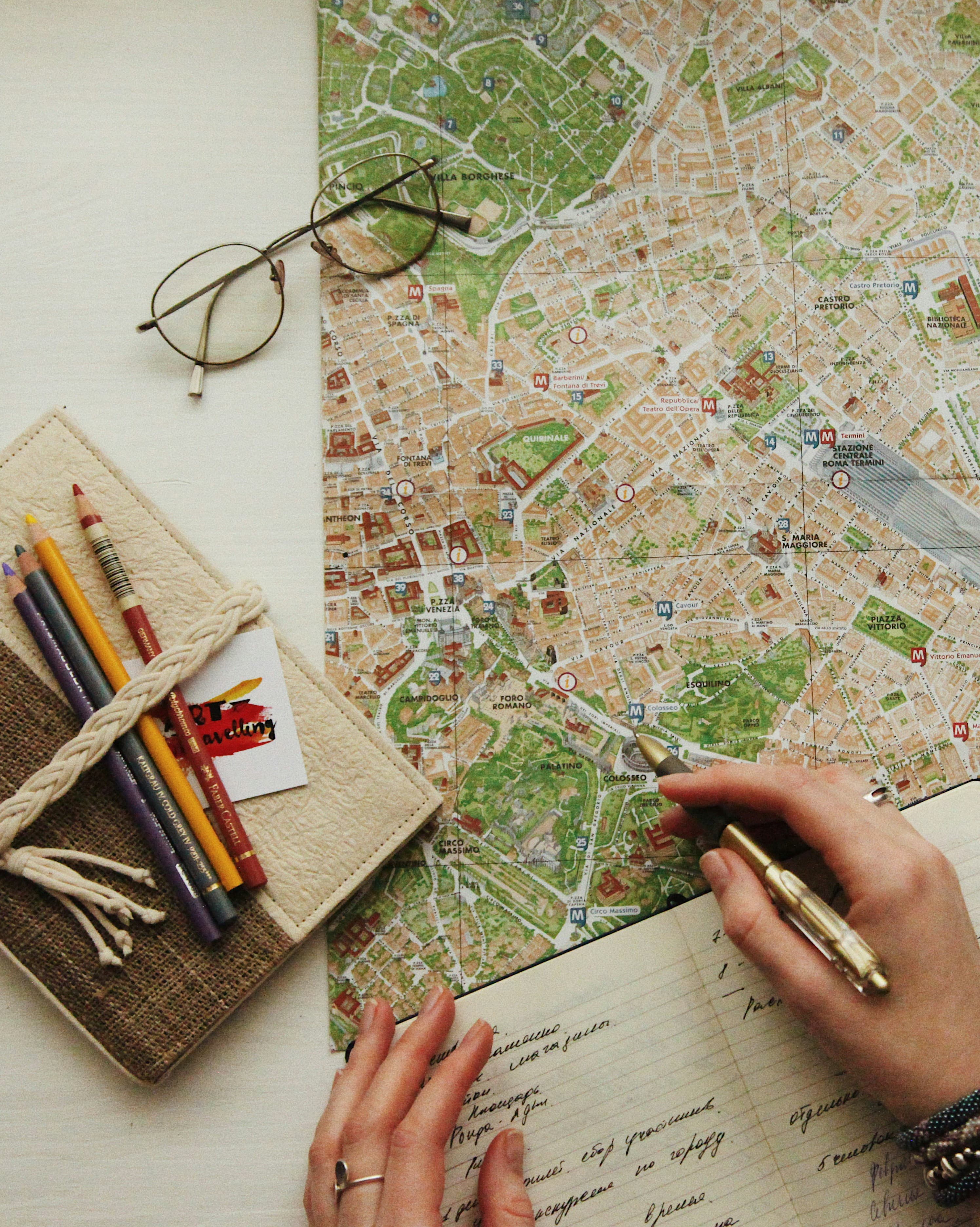 A city map marked up with a pen beside a travel journal, glasses and coloured pencils